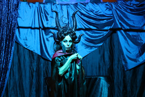 Hecate blood martini Halloween burlesque act. Witch woman with horns and moon headdress in black satin cloak preforms in Sydney Halloween cabaret show. She stands in front of blue curtain.