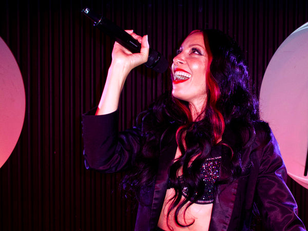 Solo singer on stage covers Cher songs from Burlesque the movie in Burlesque the Parody dinner Show. The brunette performer has  long dark hair, sequinned bra and tail coat and looks up smiling singing a big note into the hand held microphone.