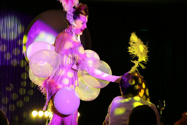 A burlesque dancer performs a traditional balloon pop routine in Talk and Tease comedy cabaret in Kings Cross. The stage lights are yellow and pink. The background is black. The burlesque dancer covered in a balloon costume, is giving a large ostrich feather to a man in the front row to pop her balloon. She is wearing a corset and feathered showgirl headdress.