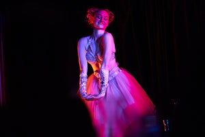 An elegant Sydney cabaret dancer performs on stage wearing a tutu She is in a pink spot light while the entire stage behind her is black so you cannot see the crowd. Her eyes are shut with a small cheeky smile and her arms stretched with her hands clasped in a girlish gesture.