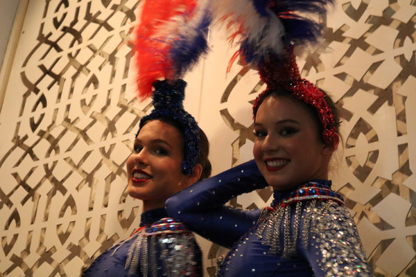 Portrait of two young French showgirls wait in the wings to perform wearing sparkling long sleeved blue dance costumes and read and blue feather headdresses. They smile at the camera in front of a cream lattice background.