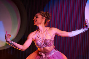 A burlesque ballet dancer performs in  in a Sydney dinner show wearing a crystal studded pink bra and pink tutu. She is doing a beautiful dance move with elegant arms and a gentle smile. Soft pink stage lighting surrounds her.