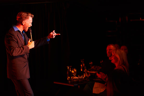 Stand up comedian and dinner show host entertains audiences at Talk and Tease Comedy Cabaret. In the amber stage lighting he stands with a microphone and wearing a dark grey suit. He motions to the crowd in a pointing gesture, mid joke. The couple on a date in the front row look with anticipation. Their champagne flutes sparkle in the ambient lighting.