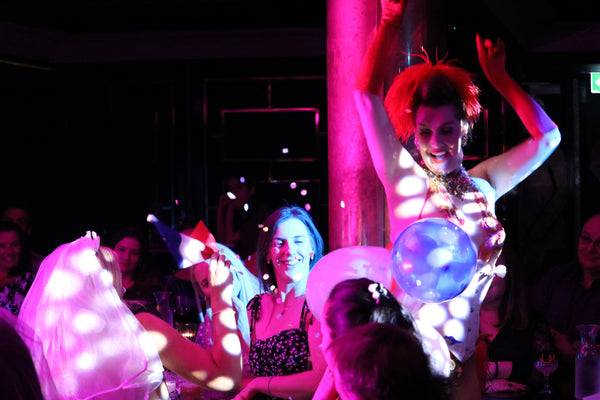 Burlesque showgirl dancers in crowd, moving between seated audience members. Her arms are in the air with one red glove. Glitter is flying everywhere from a popped balloon. Crowd are smiling as they watch the performance. Soft pink light illuminates the scene/ 