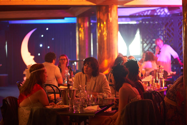 A group of women sit at a table in a daytime cabaret show lunch event. A gorgeous paper moon photo booth is in the background as light pours in from the window. The women are dressed in vintage outfits