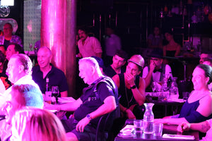 A crowd of people in a work function watch the cabaret show. They sit at their tables laughing in disbelief and taken aback. On man in the centre has his hand over his mouth. They re in a nice bar setting. 