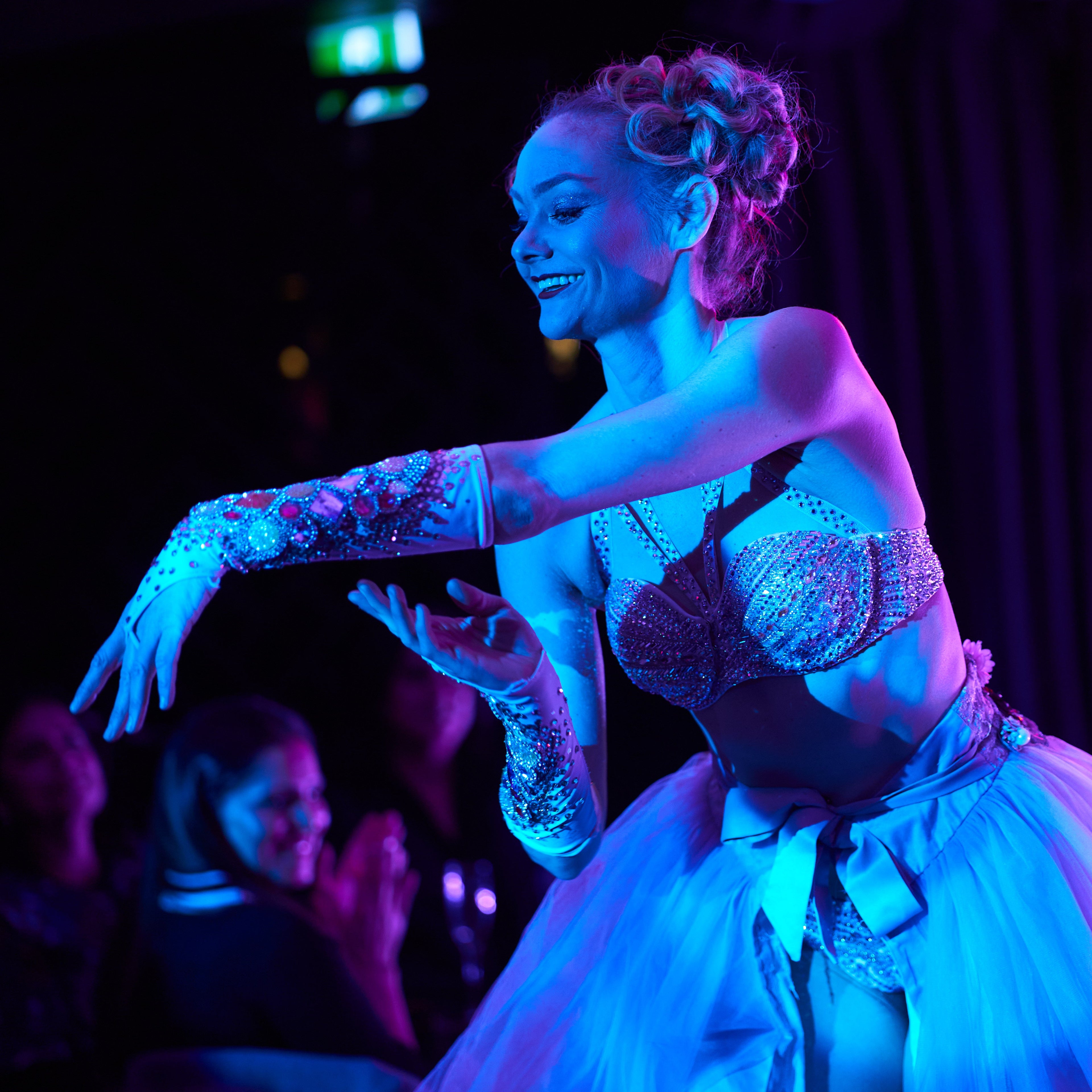 Burlesque ballet dance in tutu and bejewelled bra under blue light. Smiling as she removes a sparkling glove. 