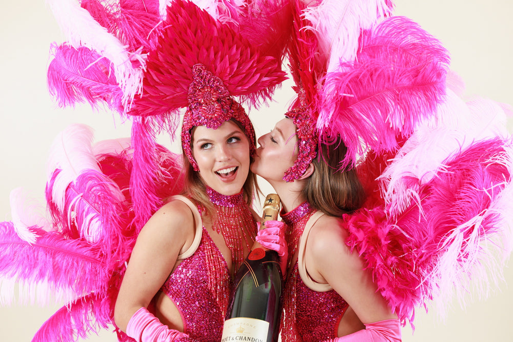 Two showgirls in bright pink feathers stand together with a jumbo bottle of Moet Chandon. One is kissing the other on the cheek in a sweet playful manner. 