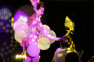 A burlesque dancer performs a traditional balloon pop routine in Talk and Tease comedy cabaret in Kings Cross. The stage lights are yellow and pink. The background is black. The burlesque dancer covered in a balloon costume, is giving a large ostrich feather to a man in the front row to pop her balloon. She is wearing a corset and feathered showgirl headdress.