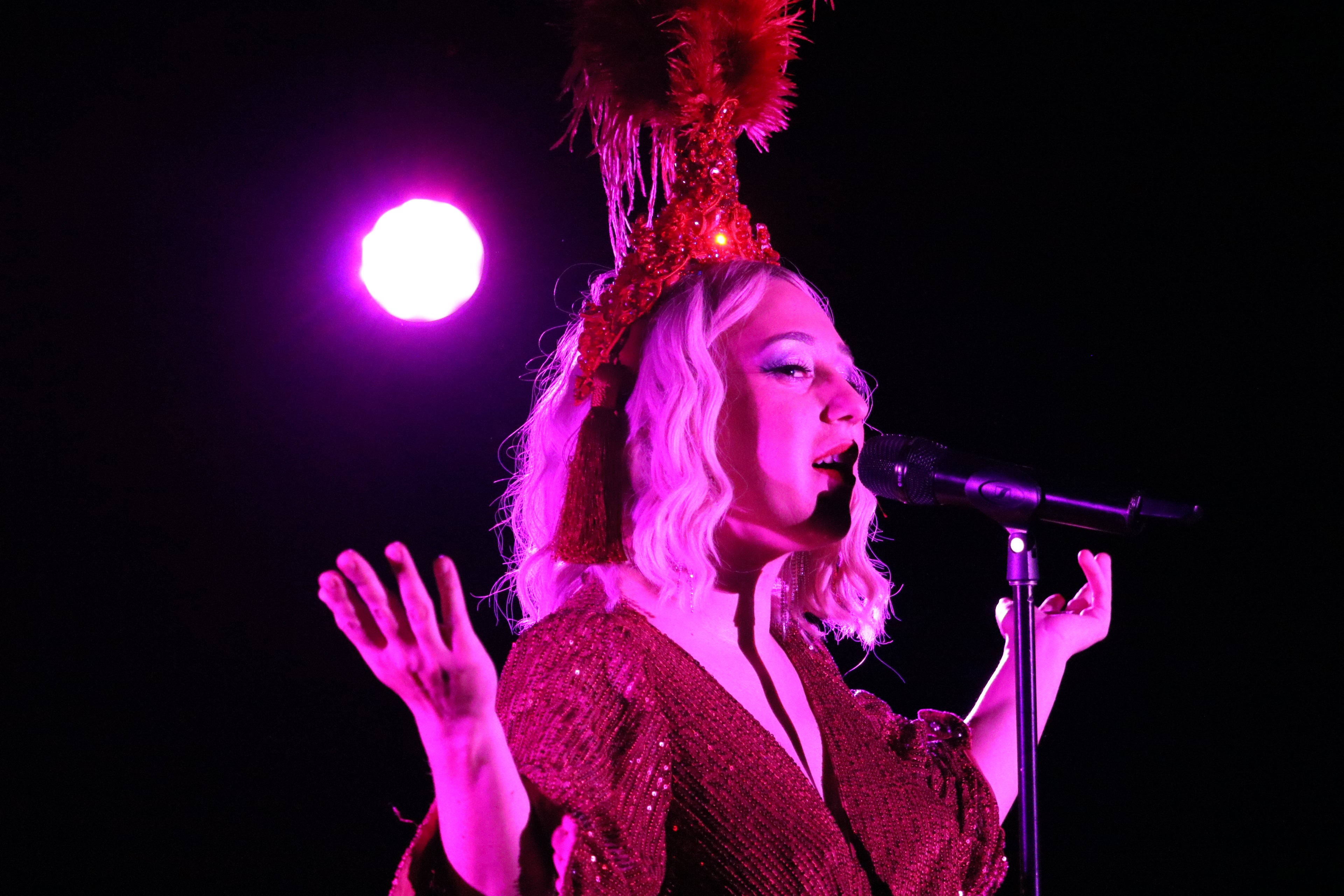A cabaret singer performs wearing a sequined red gown and red feathered headdress on her platinum blonde hair. She stands in front of the microphone hands uplifted and lips parted ready to sing. A pink spotlight washes her skin while the background is in complete darkness. 
