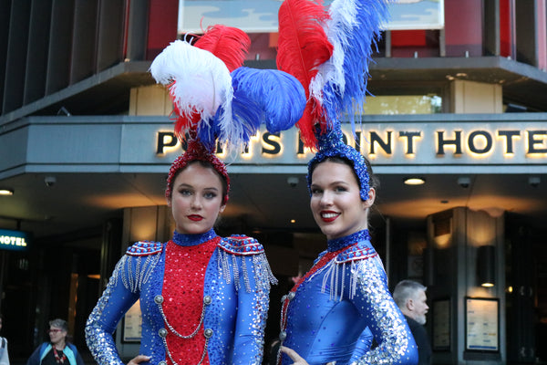 The Honeycomb Club is the place to see a traditional cabaret dinner show. Two showgirls from the Honeycomb Club, wearing feathered headpieces, stand in front of a the well-known Potts Point Hotel in Kings Cross.