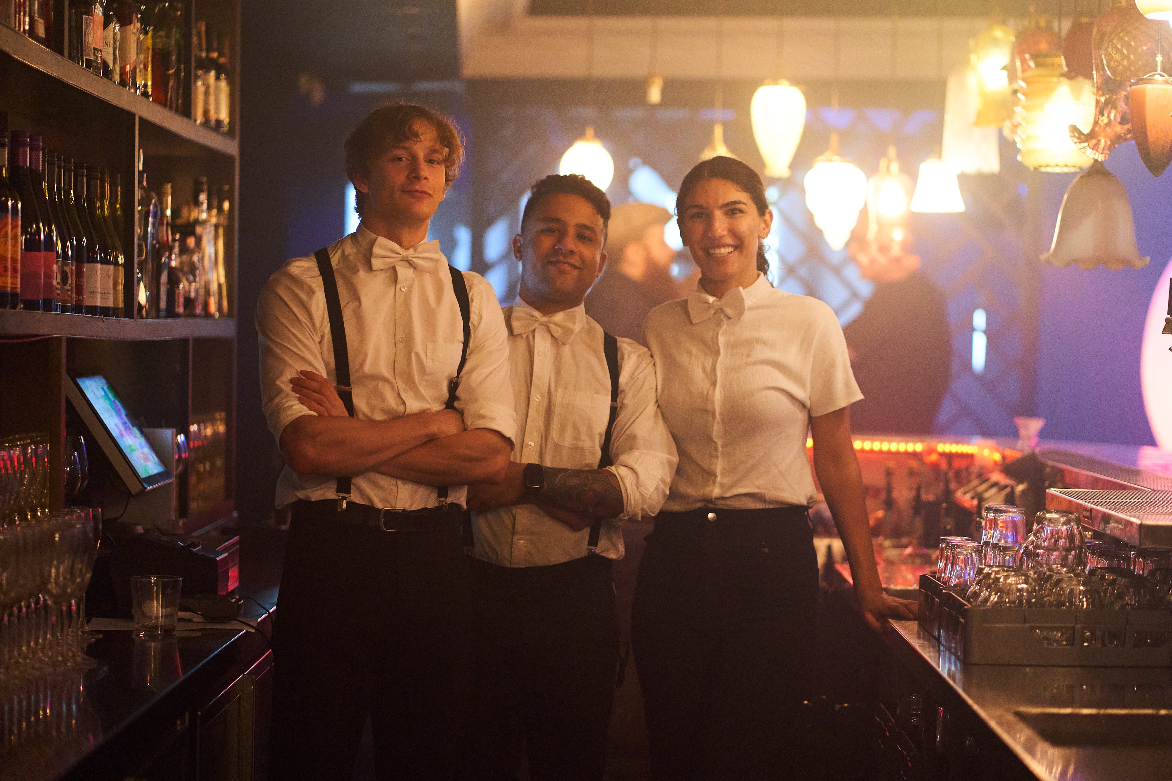 The team of waitstaff at the Honeycomb Club stand proudly shoulder to shoulder, smiling behind the bar wearing white shirts, black pants and suspenders. The ambient vintage lanterns glow above the bar in the background while the bottles and glasses glisten. 