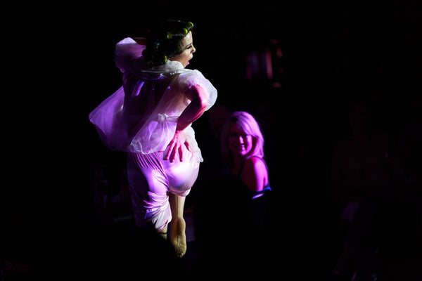 A retro burlesque dancer in an old-fashioned housewife costume strikes a dramatic pose with hand on hip and a hand behind her head as she engages the crowd that is watching from the darkness. Her vintage sleep wear is lit up in soft pink light. Her knee is lifted with her toes pointed.
