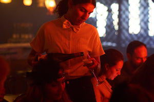 A female waiter in a bow tie serves sparkling wine to a female guest seated at her cabaret table while surrounded by other patrons. The waitress hold the bottle from the bottom as she expertly pours it into a champagne flute. Sunlight and ambient lighting from the bar illuminate the background. 