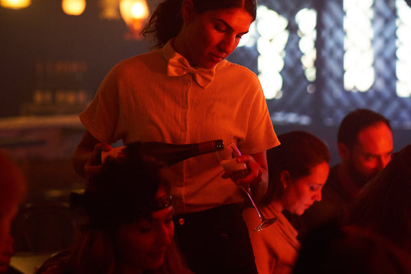 A female waiter in a bow tie serves sparkling wine to a female guest seated at her cabaret table while surrounded by other patrons. The waitress hold the bottle from the bottom as she expertly pours it into a champagne flute. Sunlight and ambient lighting from the bar illuminate the background. 